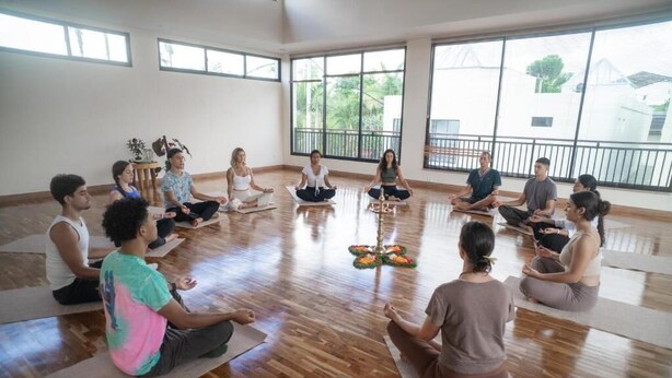 Un grupo de personas sentadas sobre tapetes de yoga en una habitación con ventanas amplias y suelo de madera.