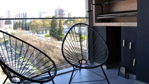 A balcony with two black wire chairs, a view of a parking lot, and a cityscape in the background.
