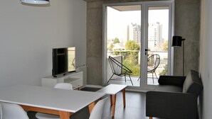 A modern living space with a flat-screen TV on a white cabinet, a dining table with chairs, a black chair, and a sofa.