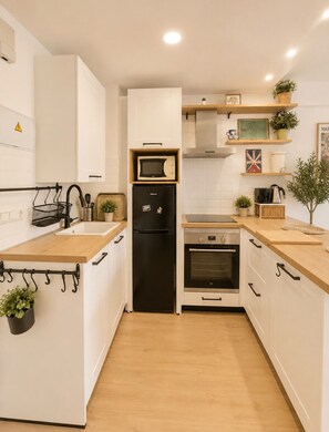 A modern kitchen with white cabinetry, a black refrigerator, a microwave, and an oven. There are wooden countertops, a sink, and various kitchen utensils. - Terrasol Berlin 2 in Torre del Mar (Velez-Malaga)