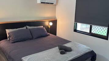 A bedroom with a bed, a bedside lamp, an air conditioning unit, and a window with a blind.