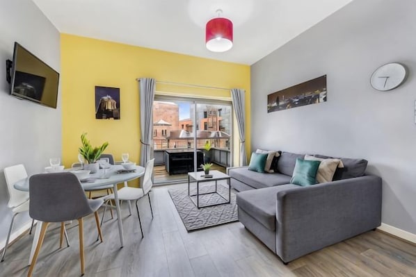 A modern living room with a grey sofa, a dining table with chairs, a coffee table, and a TV mounted on the wall.