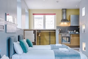 A modern hotel room with a bed, a small kitchenette, and a view of the city.