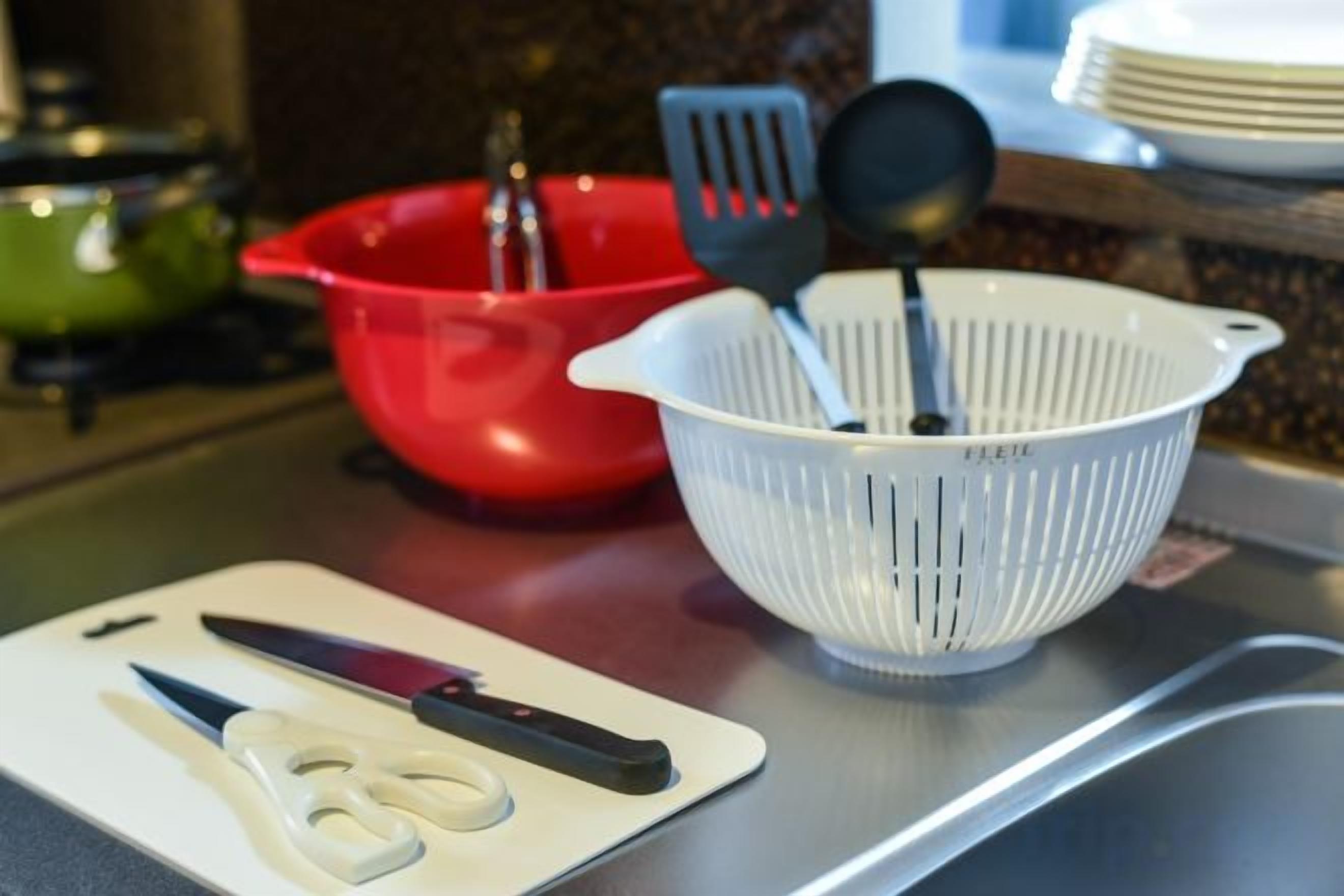 A kitchen counter with a white cutting board, a pair of scissors, a red bowl, and a white colander with utensils.