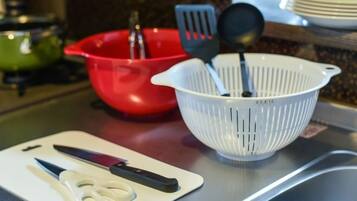 A kitchen counter with a white cutting board, a pair of scissors, a red bowl, and a white colander with utensils.