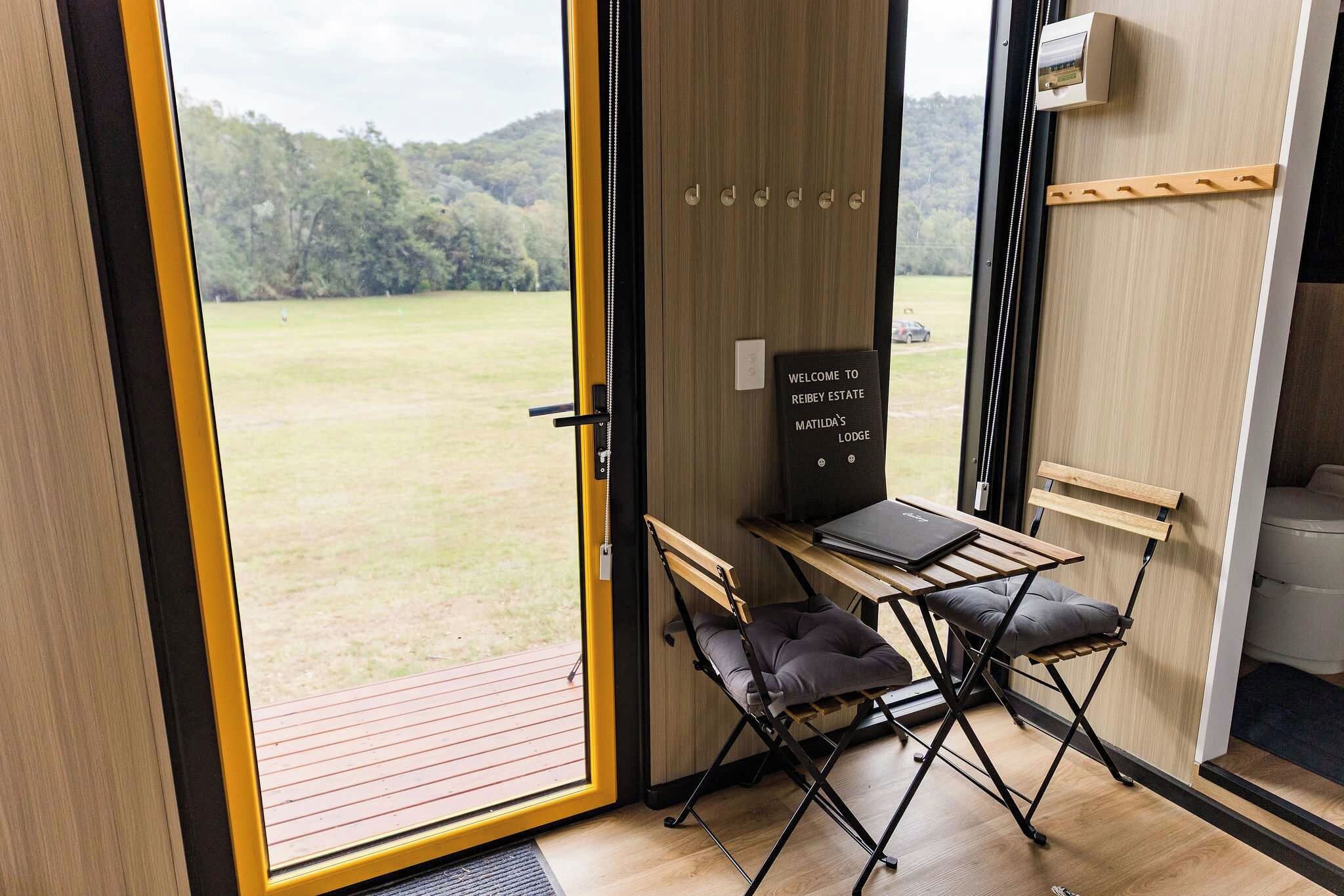 Een kamer met een houten vloer, een klein tafeltje met een stoel, een bordje met de tekst 'Welcome to Reyney Estate Matalida's Lodge' en uitzicht op een grasveld door een groot raam.
