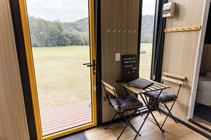A room with a wooden floor, a small table with a chair, a sign reading 'Welcome to Reyney Estate Matalida's Lodge', and a view of a grassy field through a large window.