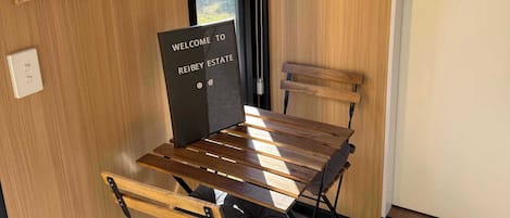 A room with a wooden table, chairs, a hanging coat rack, a welcome sign, and a small electronic device on the wall.