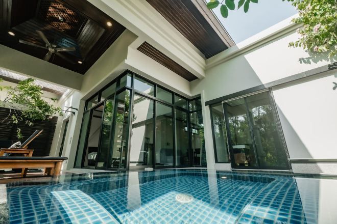 A modern indoor swimming pool with glass walls, a ceiling fan, and a wooden bench.