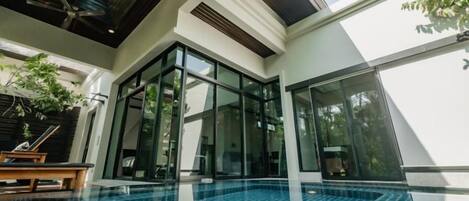 A modern indoor swimming pool with glass walls, a ceiling fan, and a wooden bench.