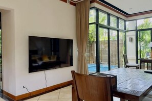 A room with a flat-screen TV mounted on the wall, a dining table with chairs, and large windows offering a view of the outdoors. - Rose Pool Villa, The perfect private oasis (Phuket)