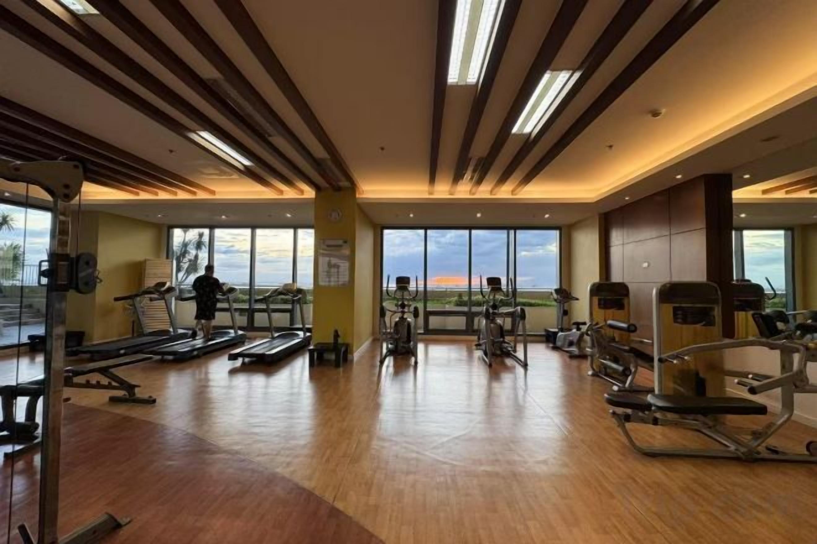 A gym with exercise equipment, large windows, and a wooden floor.