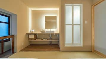 A room with a bed, a bedside table, a mirror, a window, and a bathroom with a sink and mirror.
