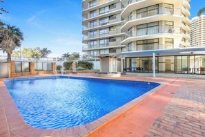 A swimming pool with a brick border, surrounded by a building with multiple balconies and a covered area. - The Ocean Arcade - Views, Games & Vibes (Surfers Paradise)