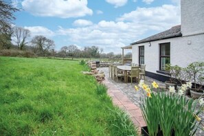 A house with a paved patio, outdoor seating, and a garden with daffodils. - Hill House - 4 Bedroom Holiday Home - Llanrhidian (Llanrhidian Lower)