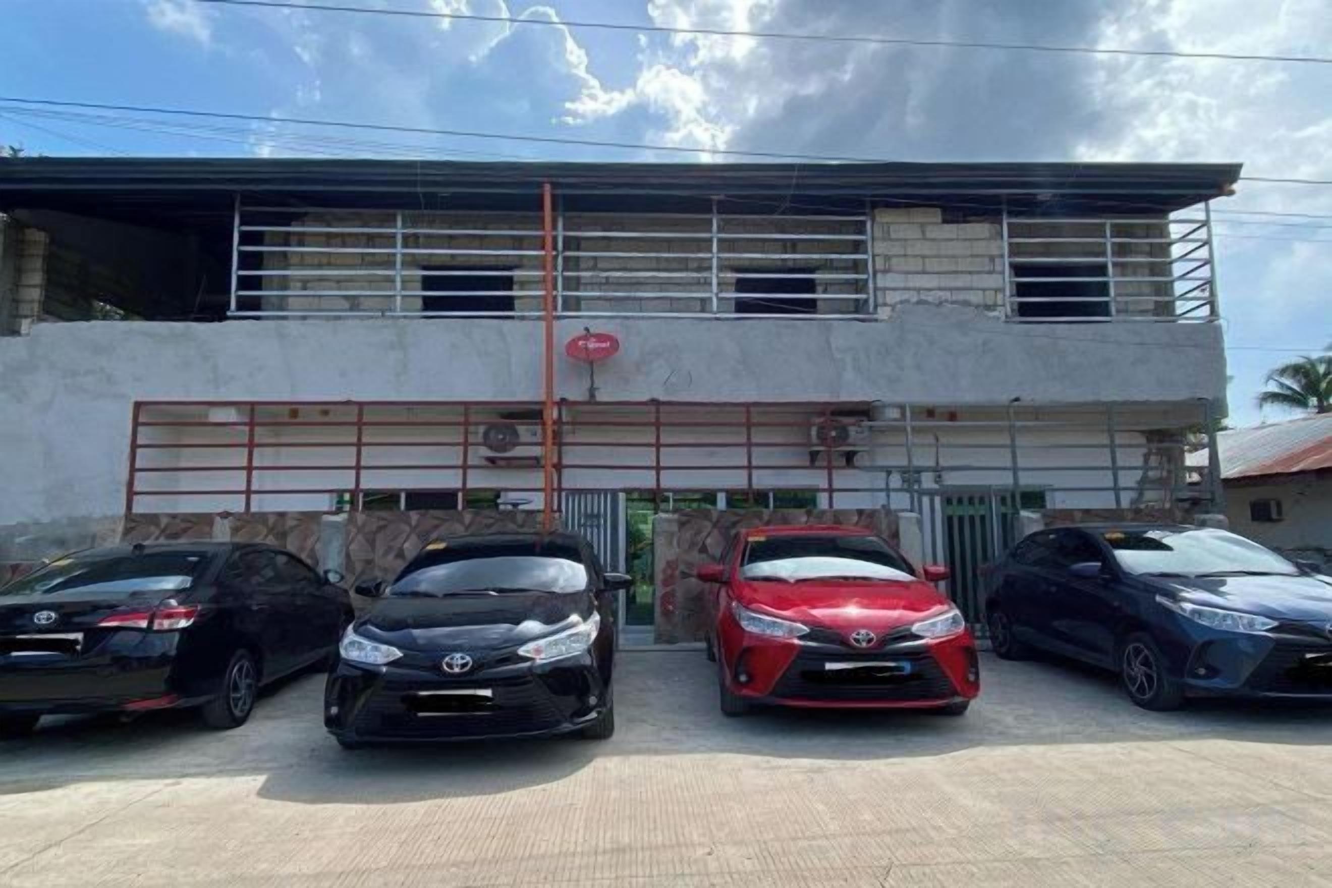 Un edificio con entrada protegida por una reja, tres coches estacionados y cielo despejado.