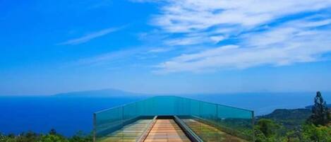 A glass bridge leading to a high vantage point with a view of the sea and distant hills.