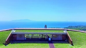 A modern building with a glass facade, situated on a grassy hill overlooking a coastal view.