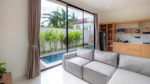 A modern living room with a sofa, a kitchen area with cabinets and appliances, and a view of a swimming pool.