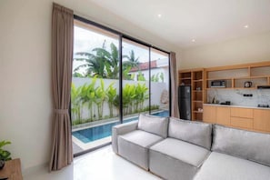A modern living room with a sofa, a kitchen area with cabinets and appliances, and a view of a swimming pool. - Villa Sardegna Ubud (Bali)