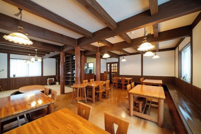 Interior de restaurante con mesas y sillas de madera, techos altos con vigas a la vista e iluminación colgante.