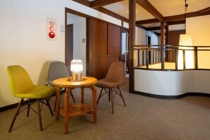 Una habitación con un banco curvo, dos sillas, una mesa con lámpara y una puerta que da a otra estancia.