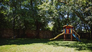 Uma área de recreação infantil com escorregador e estrutura de escalada, cercada por árvores e um muro de pedra.