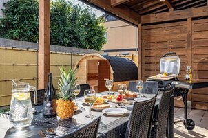 Un área de comedor al aire libre con una parrilla, sillas de mimbre y una mesa dispuesta con platos, vasos y una piña.
