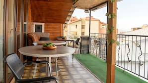 Un balcón de madera con una mesa y sillas, un pequeño jardín y vistas a edificios vecinos.