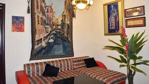 Una habitación con un cuadro grande de una escena callejera, un sofá con cojines estampados, una araña y cuadros enmarcados en la pared.