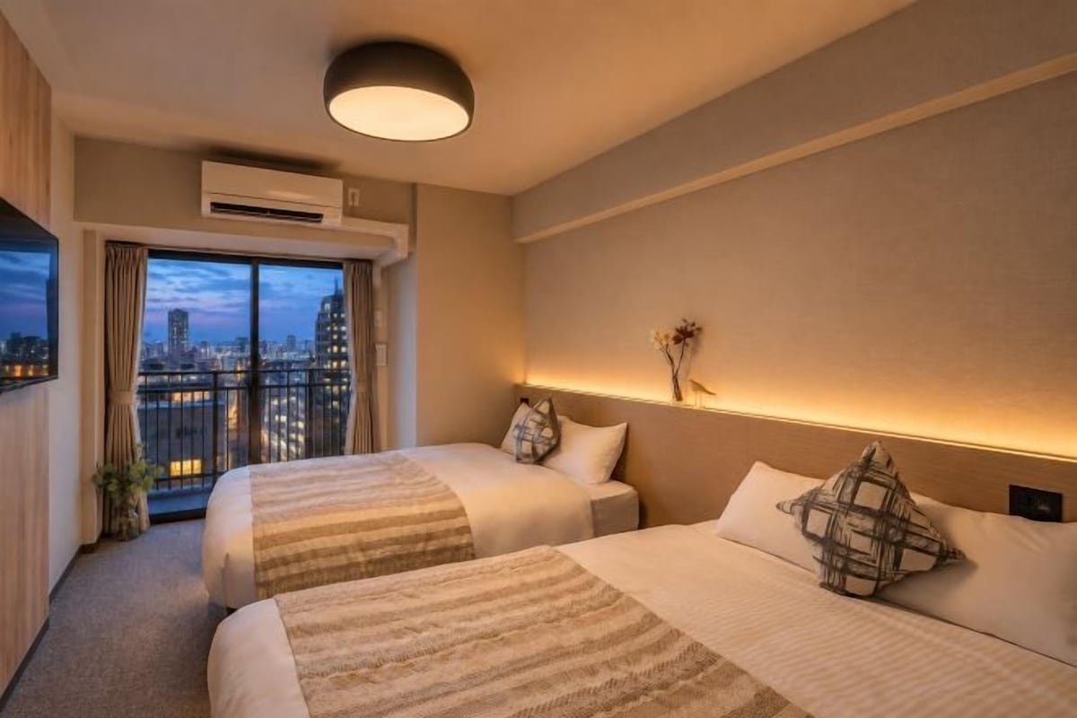 A hotel room with two beds, a city view, a bedside table with a lamp, and a wall-mounted air conditioner.