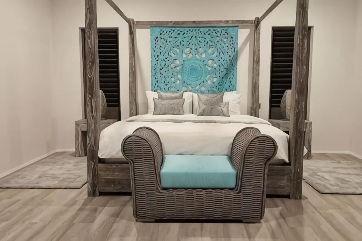 Un lit à baldaquin avec un banc en osier placé devant, une tapisserie à motifs accrochée au-dessus et un plancher en bois.