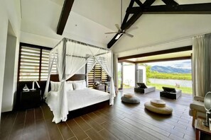 A four-poster bed with a canopy, a ceiling fan, and a view of a natural landscape through large windows. - Naisoso Island Villas - Fiji (Viti Levu)