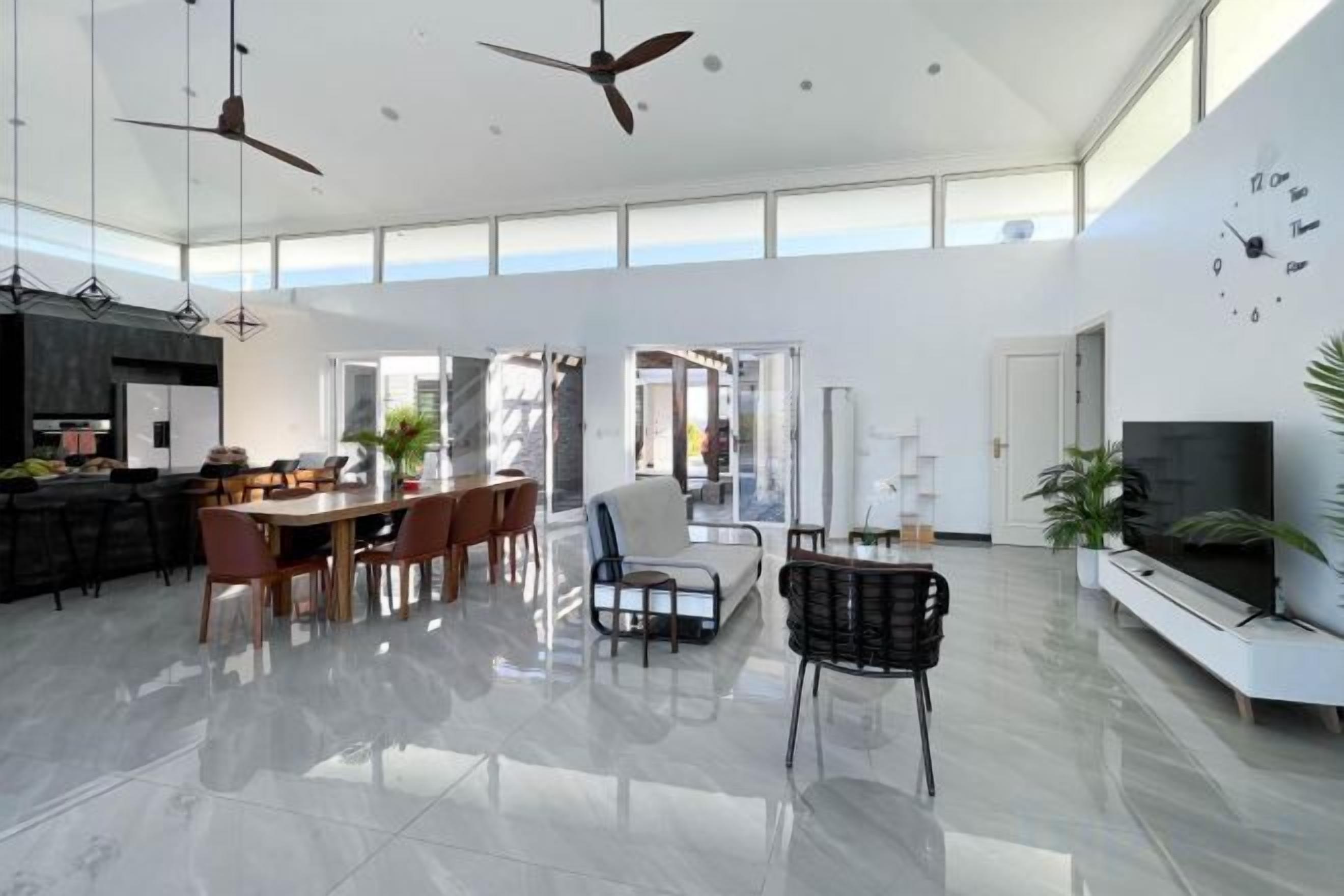 A spacious living area with a dining table, chairs, a sofa, and a TV. There are ceiling fans, a kitchen in the background, and a large window.