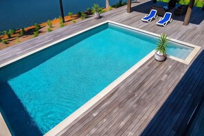 Una piscina rectangular con un deck de madera, rodeada de sillas de descanso y plantas en macetas.