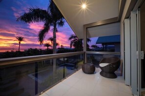 A balcony with a glass railing, two wicker chairs, and a view of a sunset over palm trees. - Naisoso Island Villas - Fiji (Viti Levu)