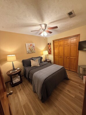 A bedroom with a bed, a nightstand, a lamp, a ceiling fan, a wardrobe, and a television. - Guest house, convenient setting, large wooded lot of towering oaks (Hollister)