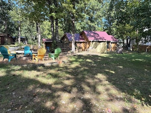 A rustic cabin with a red roof, surrounded by trees and a grassy yard. There are Adirondack chairs and a small table in the foreground. - Guest house, convenient setting, large wooded lot of towering oaks (Hollister)