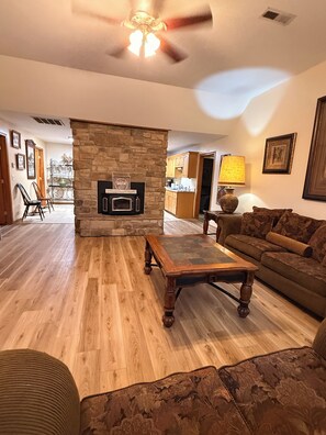 A living room with a stone fireplace, wooden flooring, a ceiling fan, and a sofa. - Guest house, convenient setting, large wooded lot of towering oaks (Hollister)