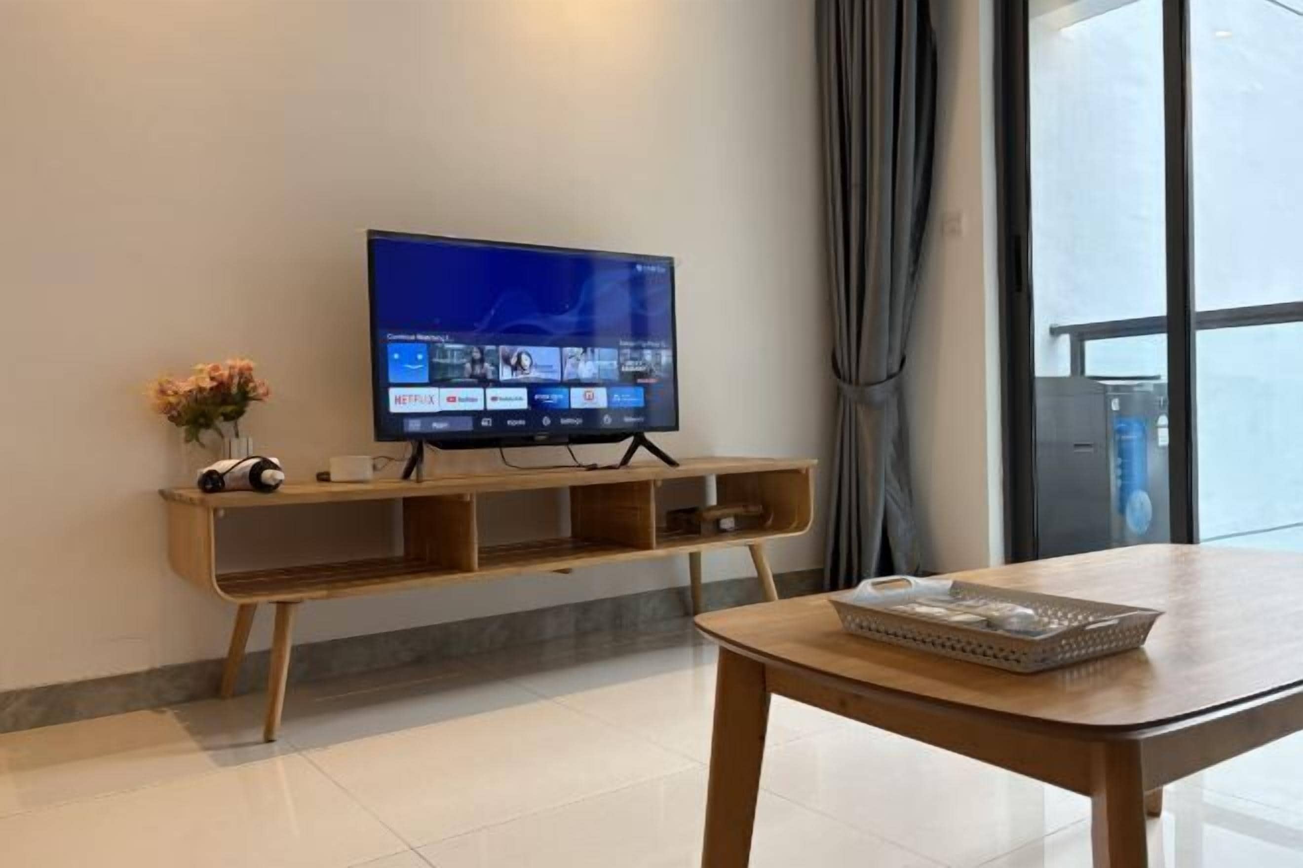 A modern living room with a flat-screen TV on a wooden console, a dining table, and a large window with curtains.