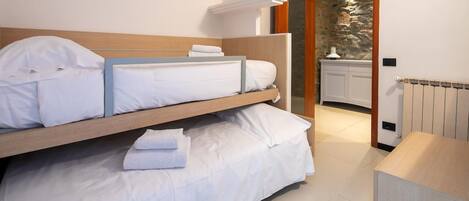 A bedroom with a bunk bed, a bedside table, a radiator, and a view of a stone wall through an open door.