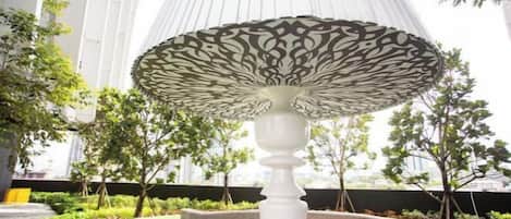 A fountain with a decorative top and a circular basin, surrounded by greenery and modern buildings.