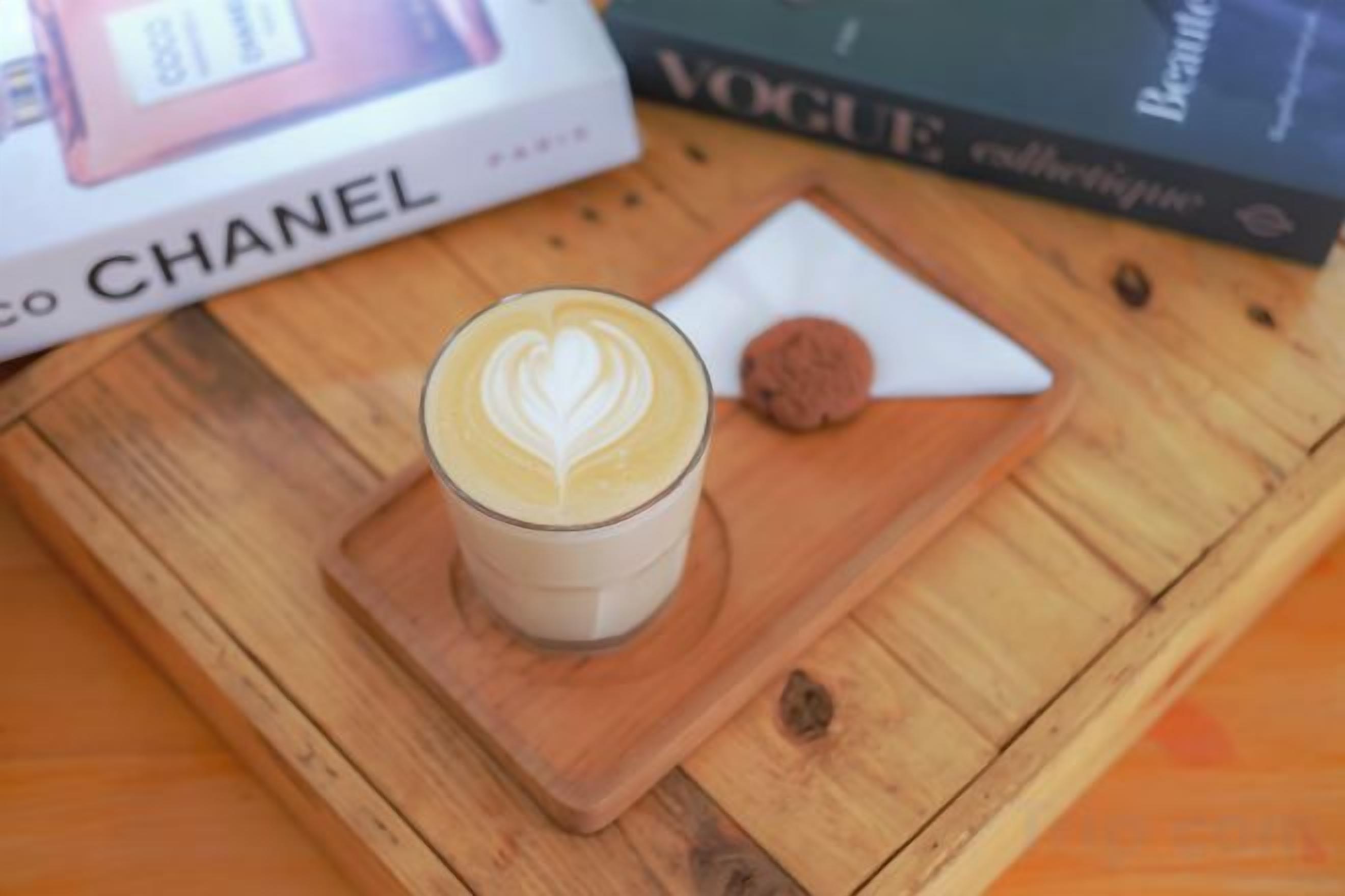 A glass of coffee with latte art on a wooden coaster, a cookie, and a book titled 'VOGUE' on a wooden table.