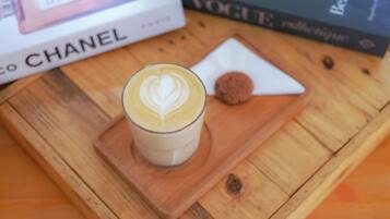 A glass of coffee with latte art on a wooden coaster, a cookie, and a book titled 'VOGUE' on a wooden table.