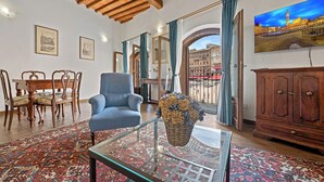 Ein Wohnzimmer mit blauem Sessel, Glastisch, gemustertem Teppich, Holzmöbeln und einem Stadtbild.