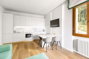 Una cocina moderna con gabinetes blancos, una mesa de comedor con sillas, un televisor de pantalla plana montado en la pared y un ventanal con vistas a zonas verdes.