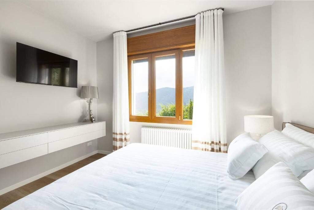 A bedroom with a bed, a TV mounted on the wall, a window with curtains, and a radiator.