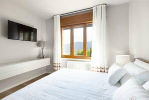 A bedroom with a bed, a TV mounted on the wall, a window with curtains, and a radiator.