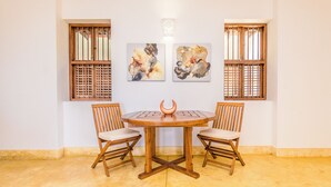 A room with a round wooden table, two chairs, and two windows with wooden shutters. There are three abstract paintings on the wall. - Cinnamon House, Weligama by The Serendipity Collection (Weligama)