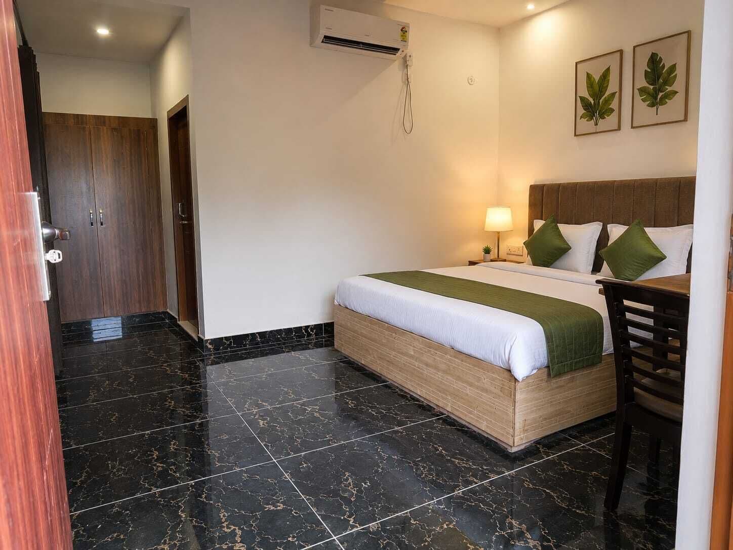 A hotel room with a bed, a desk, a chair, a nightstand, a lamp, and a wall-mounted air conditioning unit.